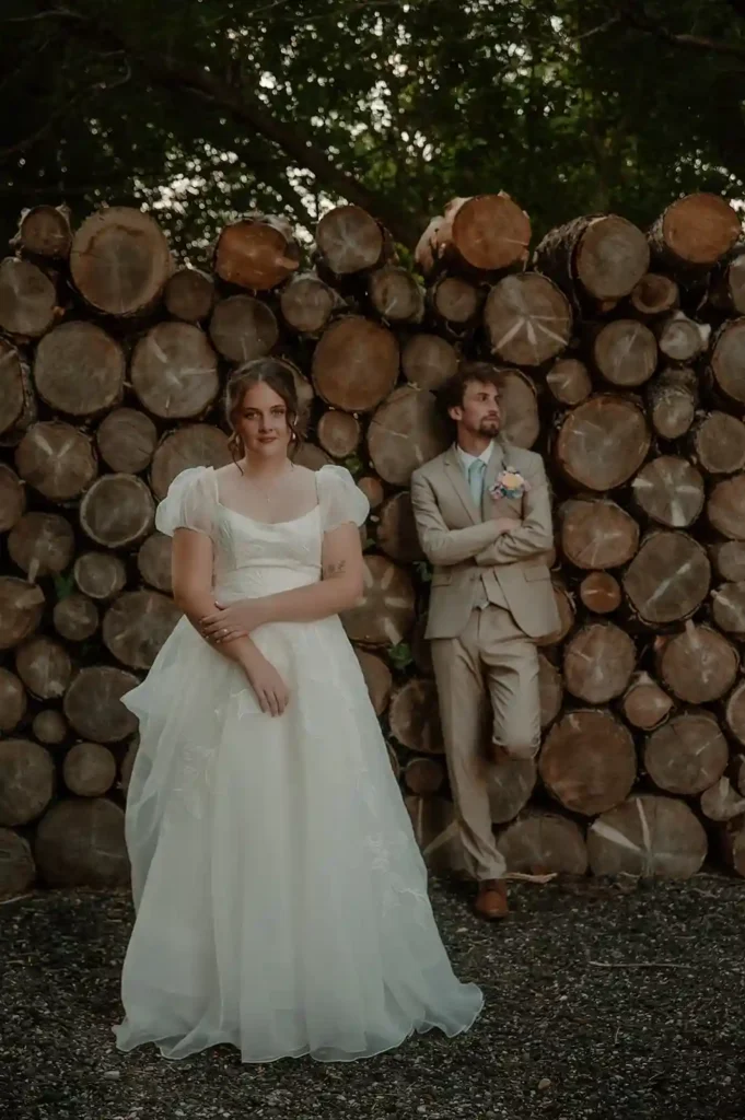 Editorial wedding portrait of bride and groom standing by a rustic woodpile
