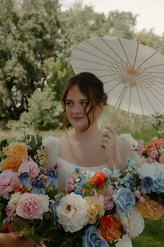 Bride holding a colorful bouquet and parasol surrounded by lush greenery