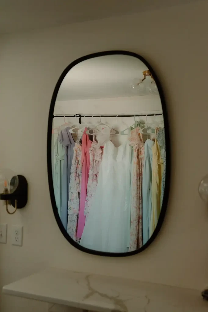 Bridesmaid dresses in pastel and floral hues hang beside the wedding gown, reflected in the mirror